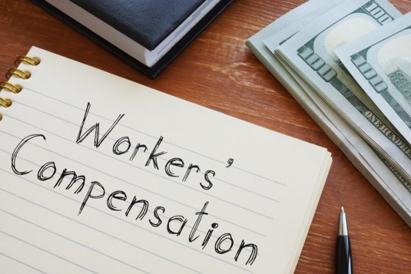 Workers' compensation written on a notepad that's on a desk with a pile of one hundred dollar bills.