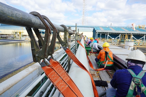 Construction workers wearing safety harnesses secured with orange straps while working on scaffolding near an industrial site.