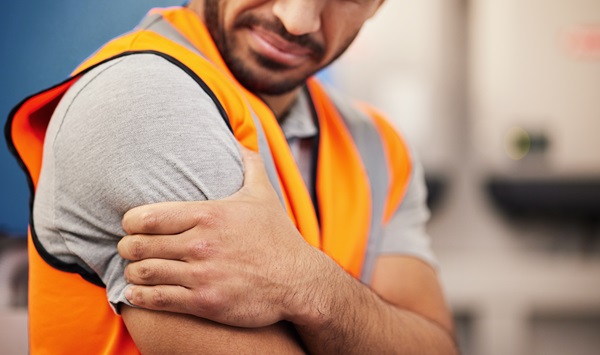 A man in an orange safety vest clutches his upper arm and shoulder, grimacing in pain from a potential work injury.