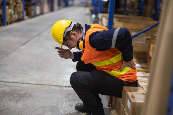 A worker in a warehouse wearing a yellow hard hat and an orange high-visibility vest is shown crouching and clutching their lower back in pain.
